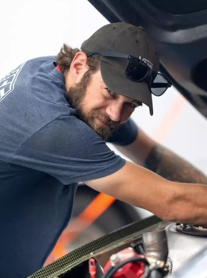A man wearing a hat and sunglasses is working under the hood of a car.