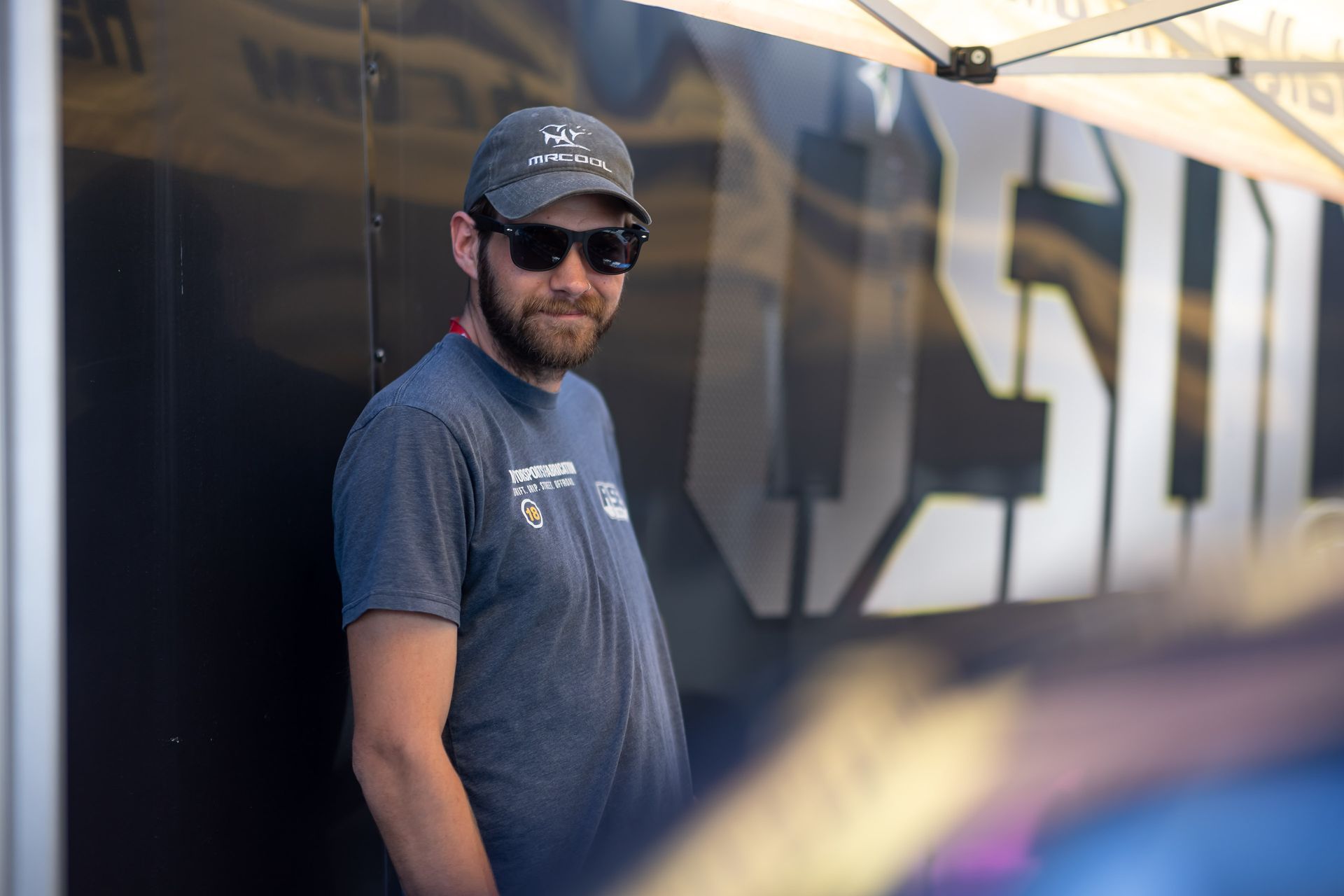 A man wearing sunglasses and a hat is standing in front of a tent.