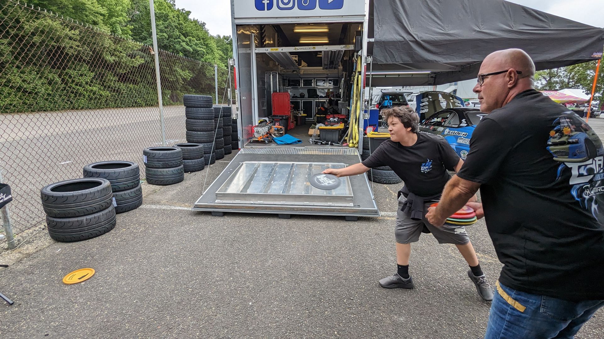 A man and a boy are playing frisbee in front of a trailer filled with tires.