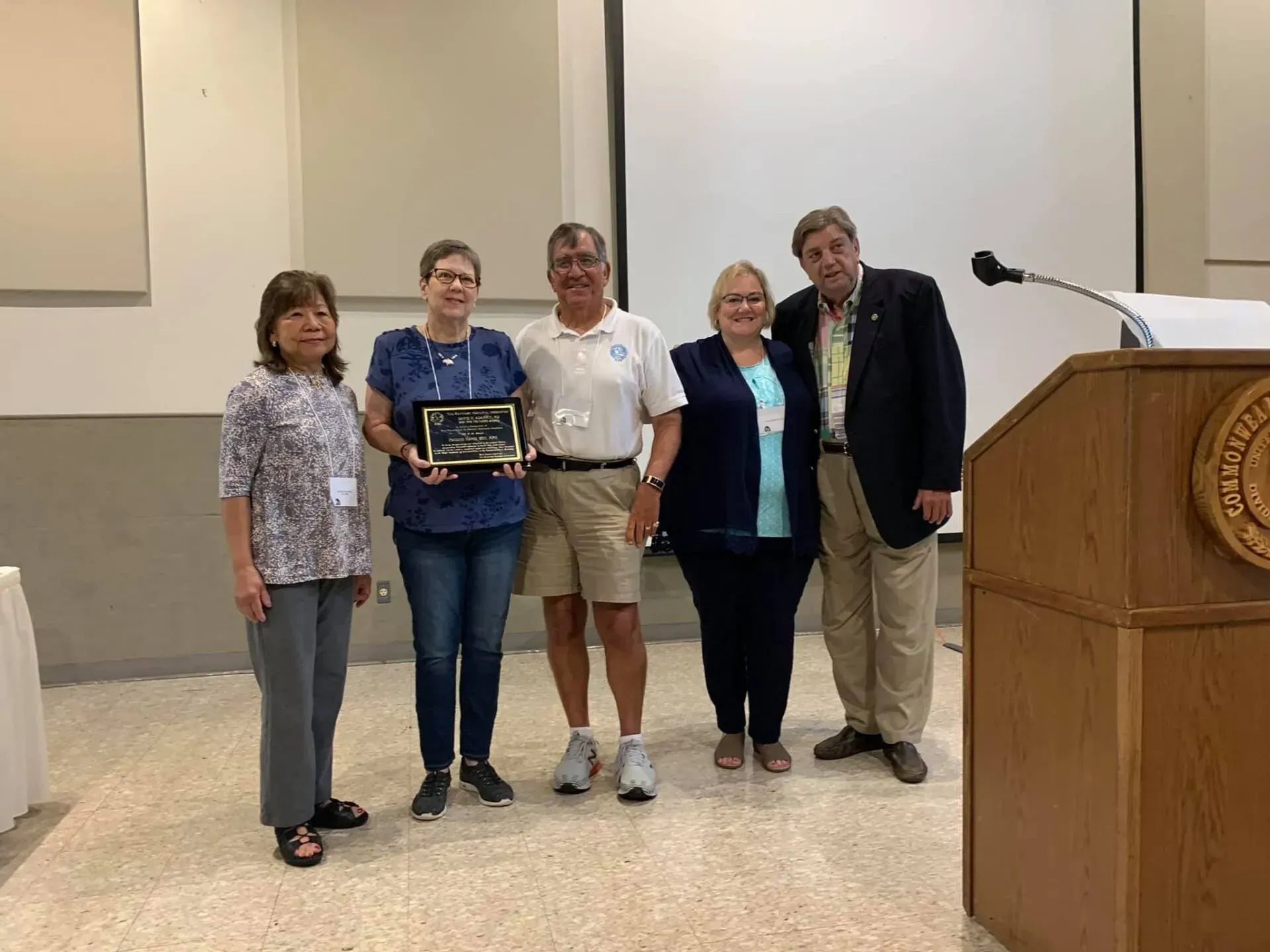 Five people pose with an award plaque near a podium. They are indoors, with a projector screen behind them.