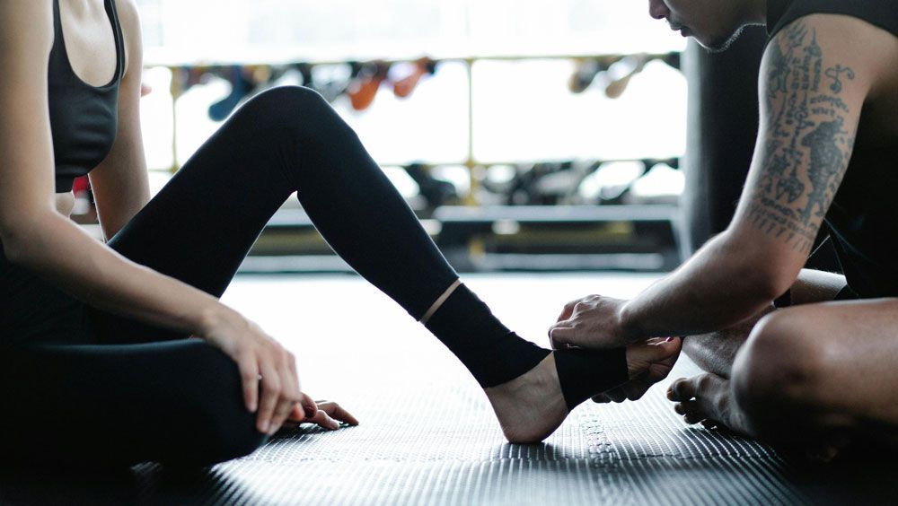 A person wraps a black athletic bandage around the ankle of another person sitting on a gym floor.
