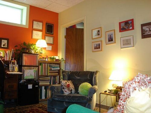 A cozy room with a dark armchair, bookshelves, and a desk against an orange wall, featuring framed art on beige walls.