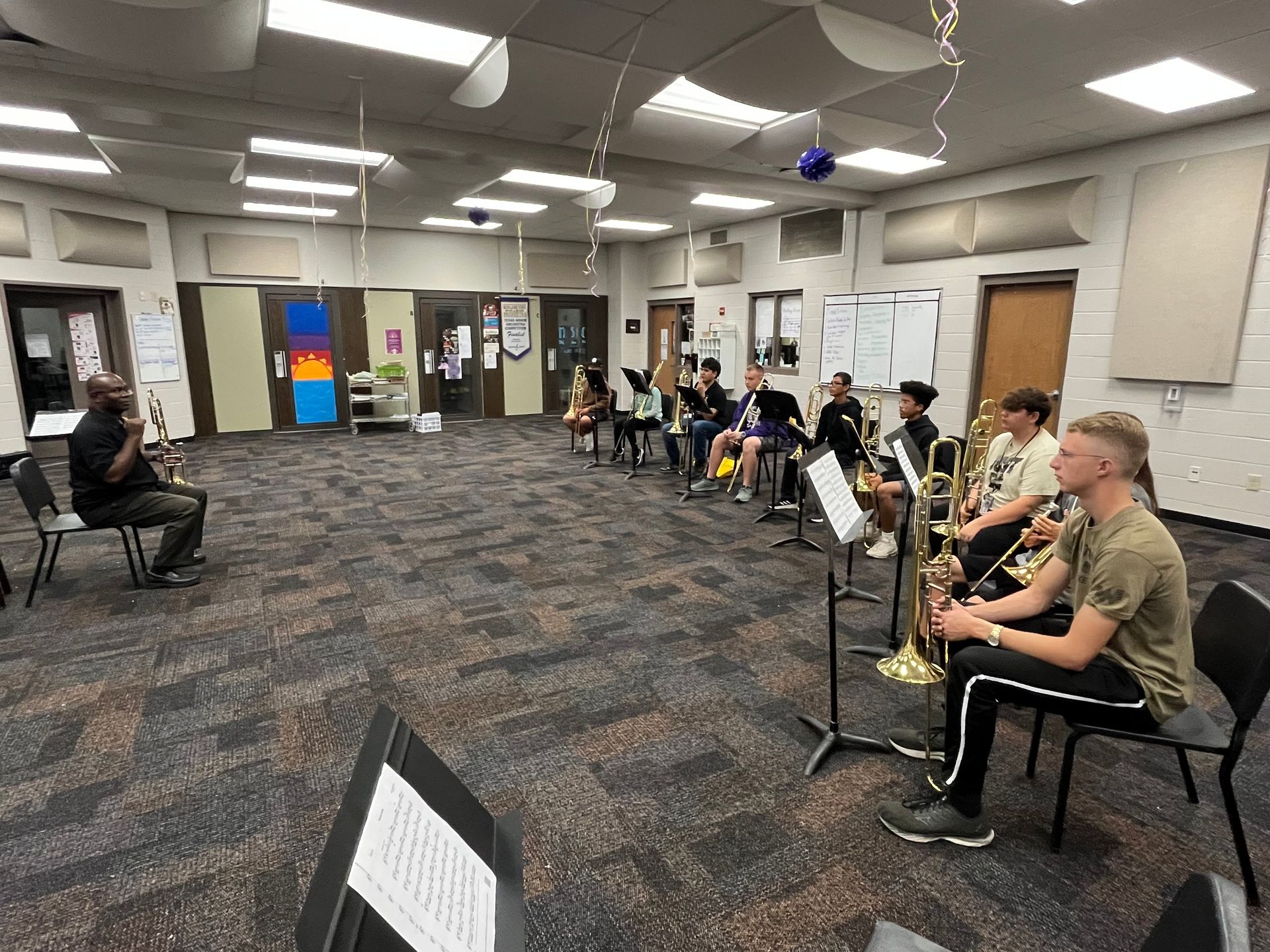 Band practice in a classroom. Students seated, some with instruments, facing a conductor.