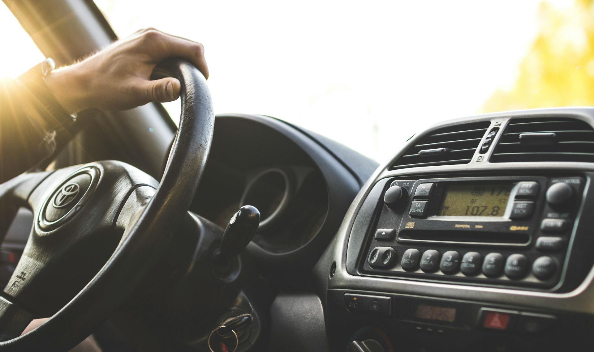 Hand on car steering wheel; dashboard with radio, vents, and sunlight.
