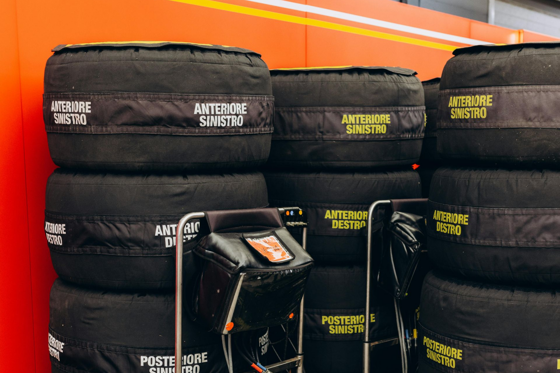 Stacks of black tires on rolling carts inside a garage, with yellow text labels. Orange and white wall in background.