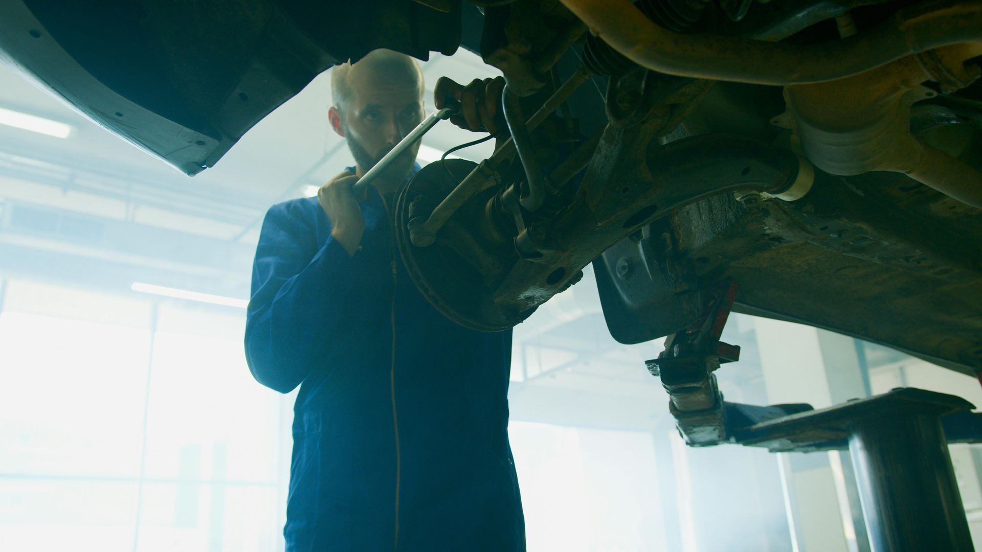 Mechanic working under a car on a lift in a bright garage, using a tool.