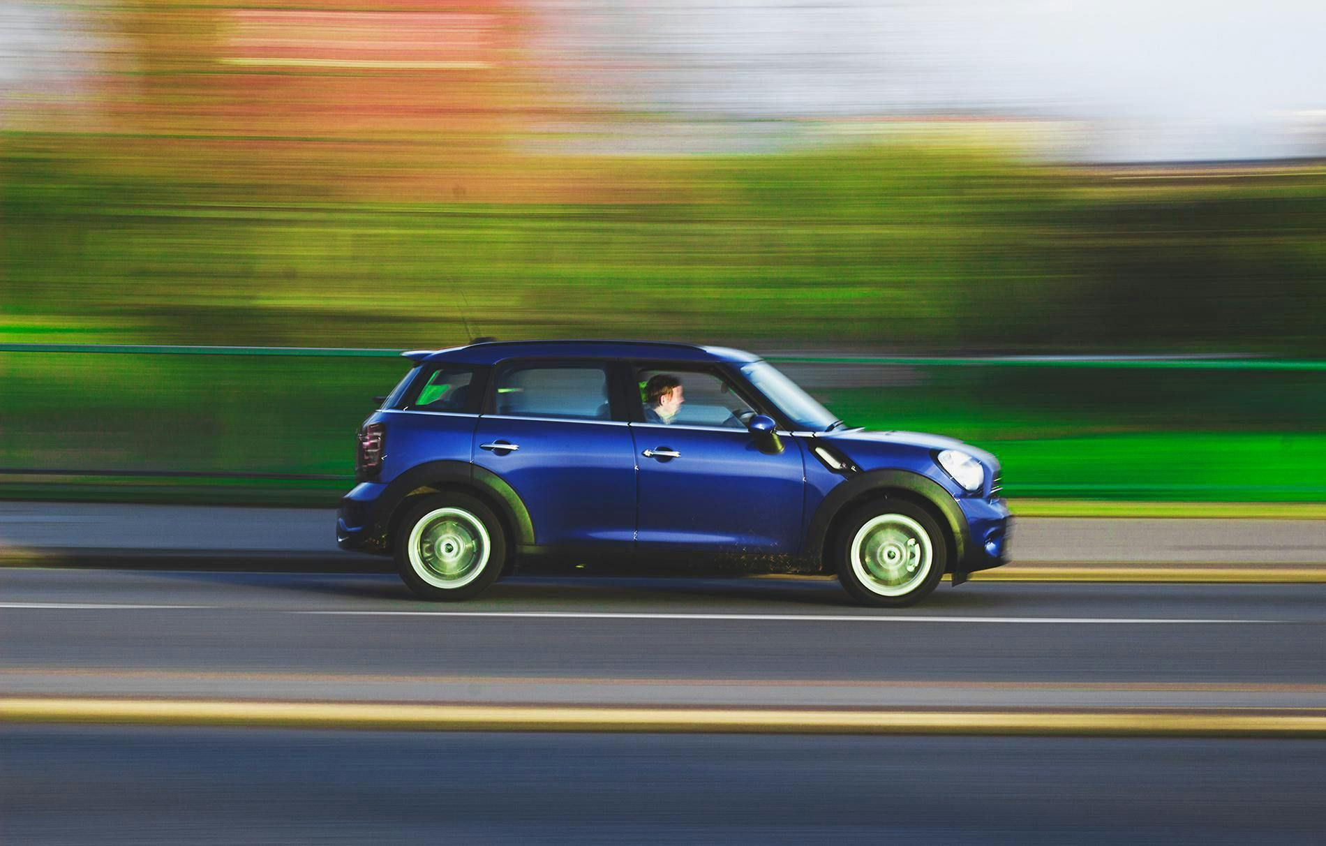 Blue Mini Cooper driving on a road, blurred background suggests speed.