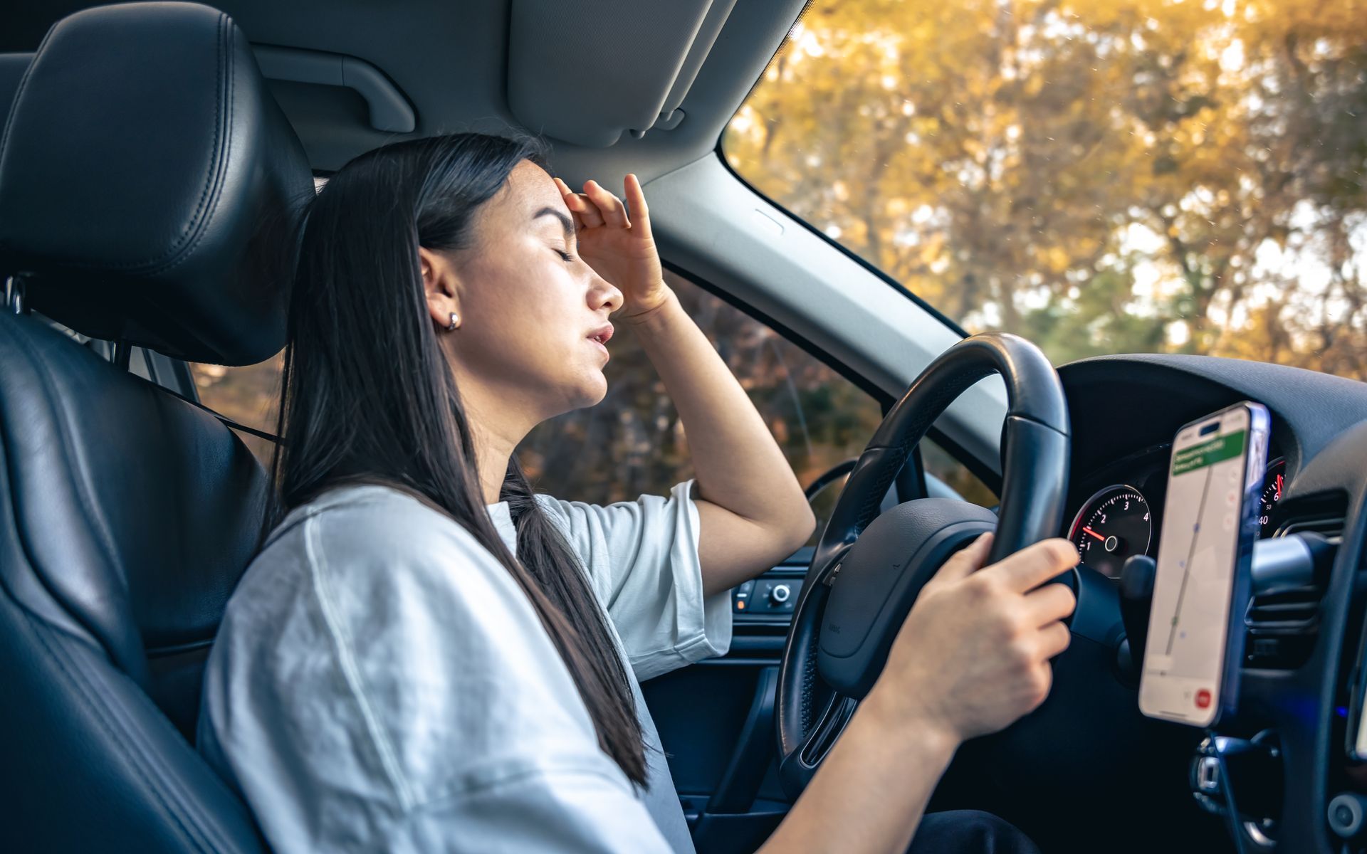 Woman in car with eyes closed, hand on forehead, phone in view; appears stressed.