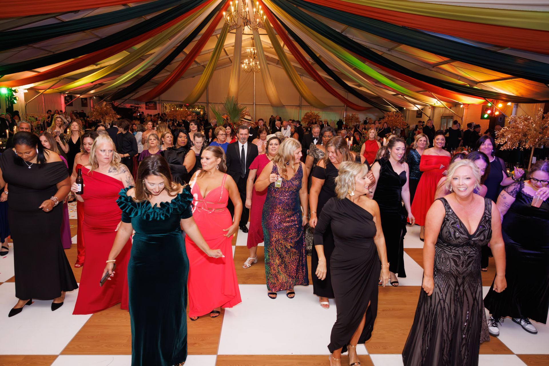 People dancing at a formal event under colorful draped tent ceiling; checkerboard floor.