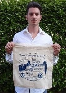 Uomo in piedi davanti al verde con in mano una borsa di tela con la grafica blu di un'auto d'epoca e una scritta in italiano.