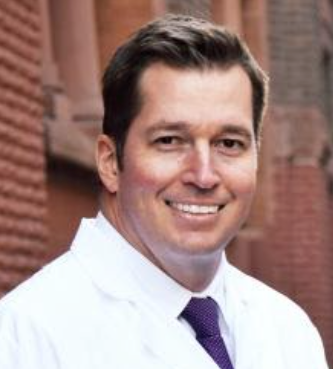 A man in a white coat and purple tie is smiling for the camera.