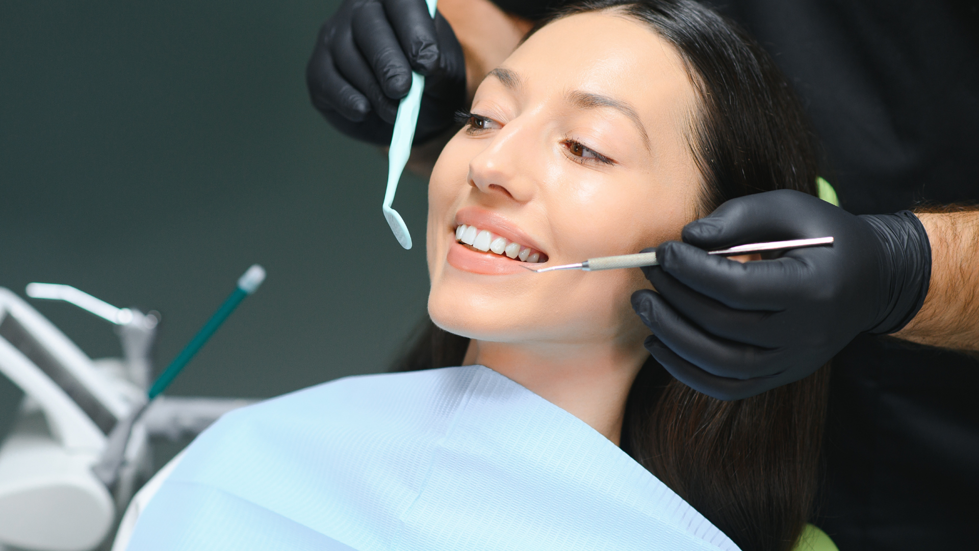 Dentist examining a patient's teeth; tools, black gloves, light blue drape, indoor setting.