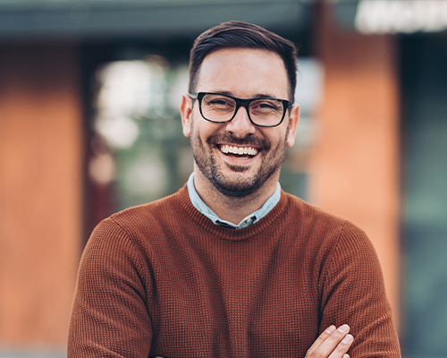 A man wearing glasses and a brown sweater is smiling with his arms crossed.