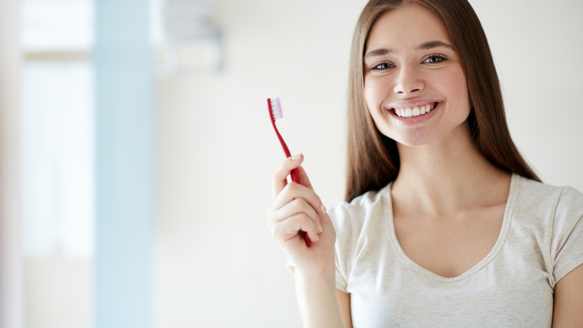 Woman smiling, holding a red toothbrush. Indoors, neutral background.