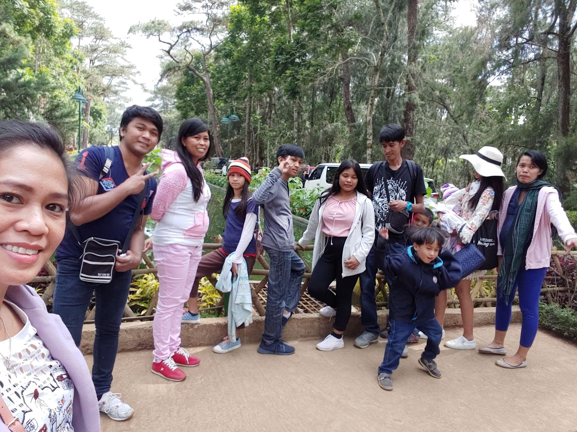 Group of people posing outdoors in a park setting. Many smiling, trees in background, sunny day.