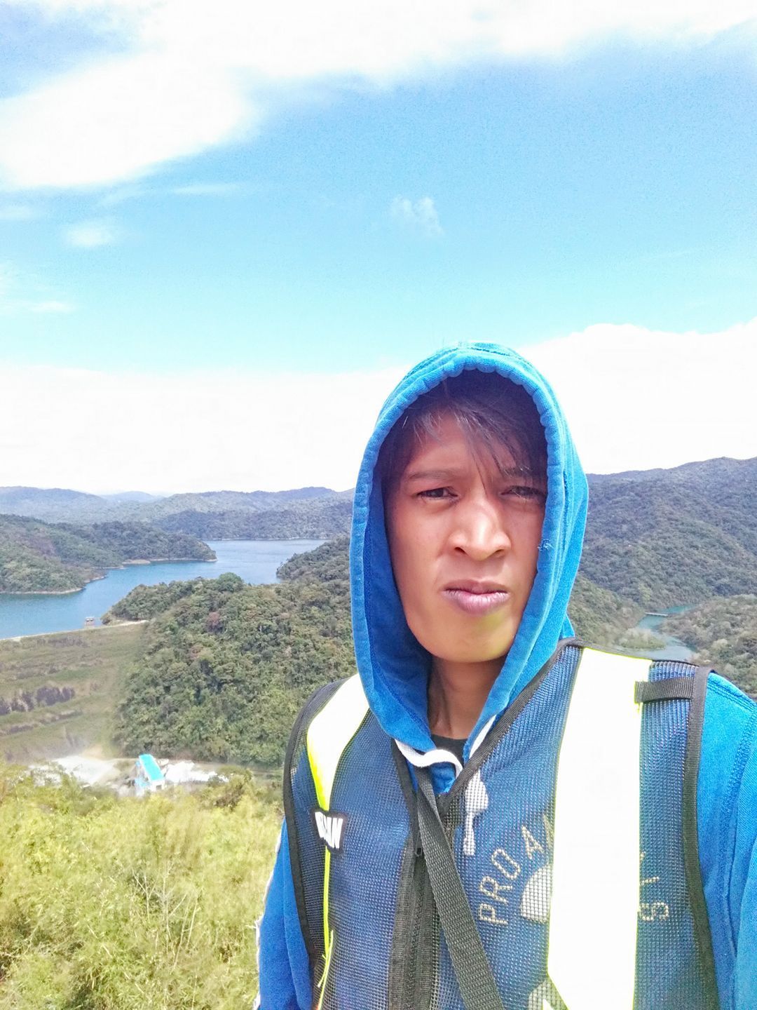 Man in blue hoodie and vest against a scenic background of a lake and hills.