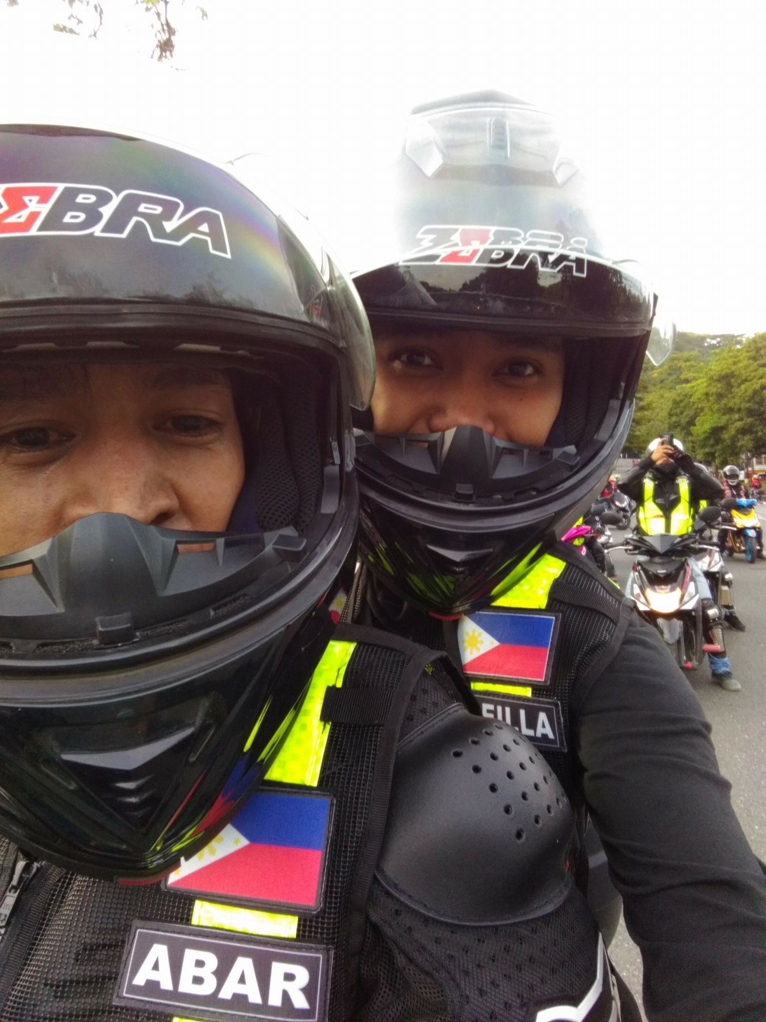 Two people on a motorcycle wearing helmets, riding with a group. They have Filipino flags on their vests.