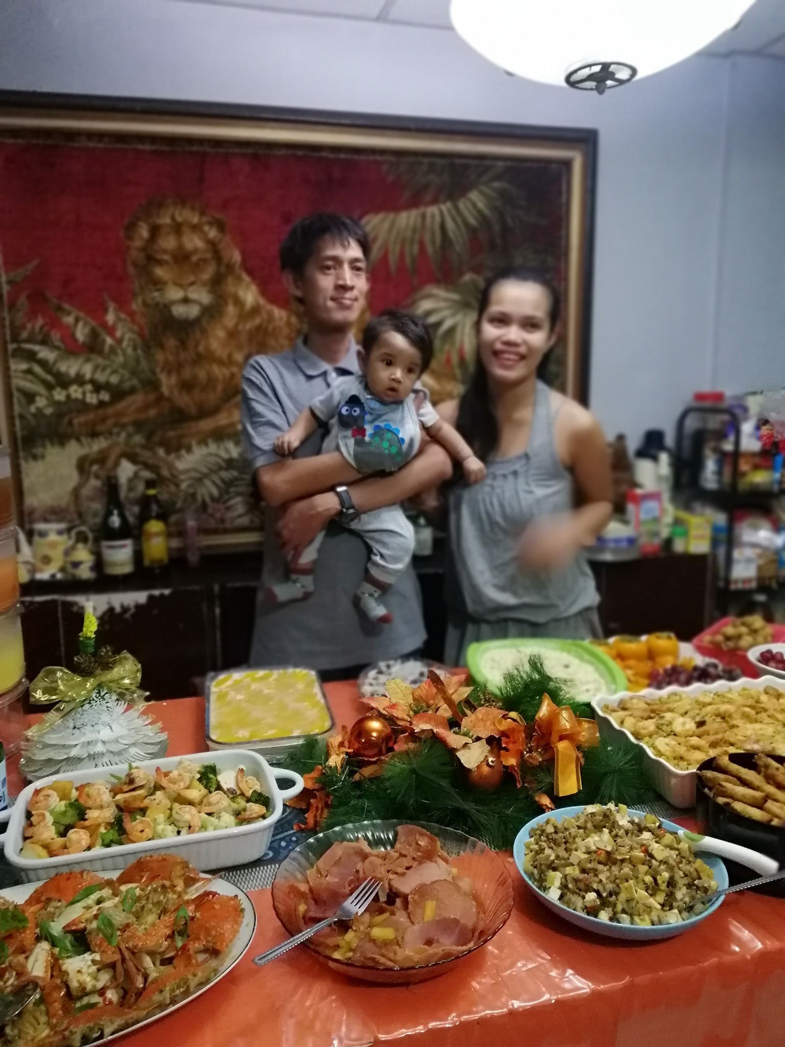 Family smiling at a table with food, including a child held by a man. Lion tapestry in the background.