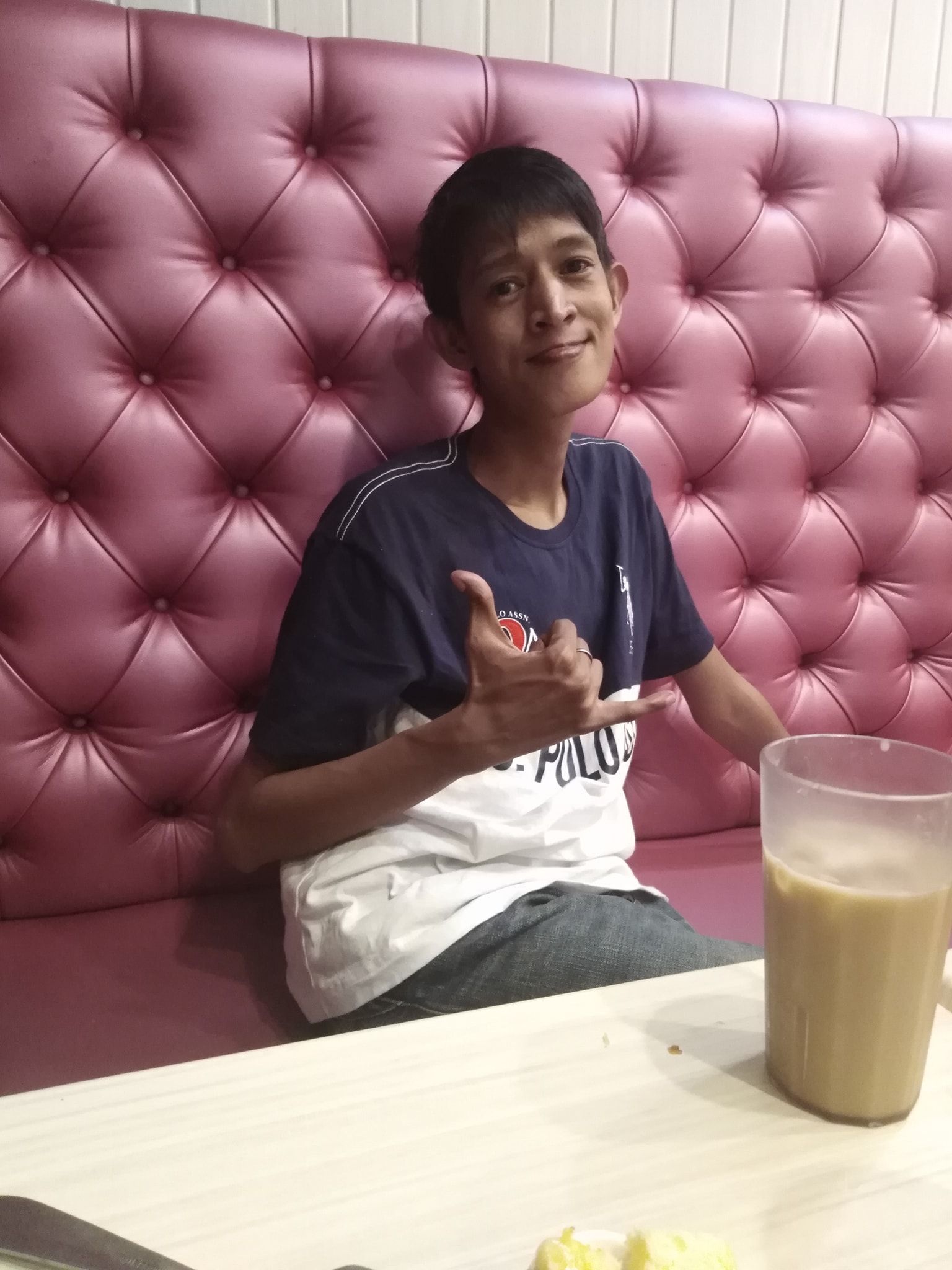 Person giving a thumbs-up gesture while seated at a table, with a drink beside them. They smile, near a pink-tufted wall.