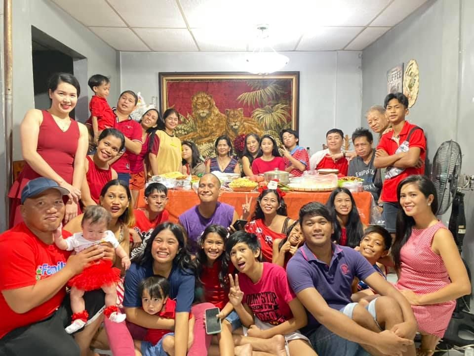 Large family gathered around a table; many wearing red. Smiling, in a brightly lit room with a painting on the wall.