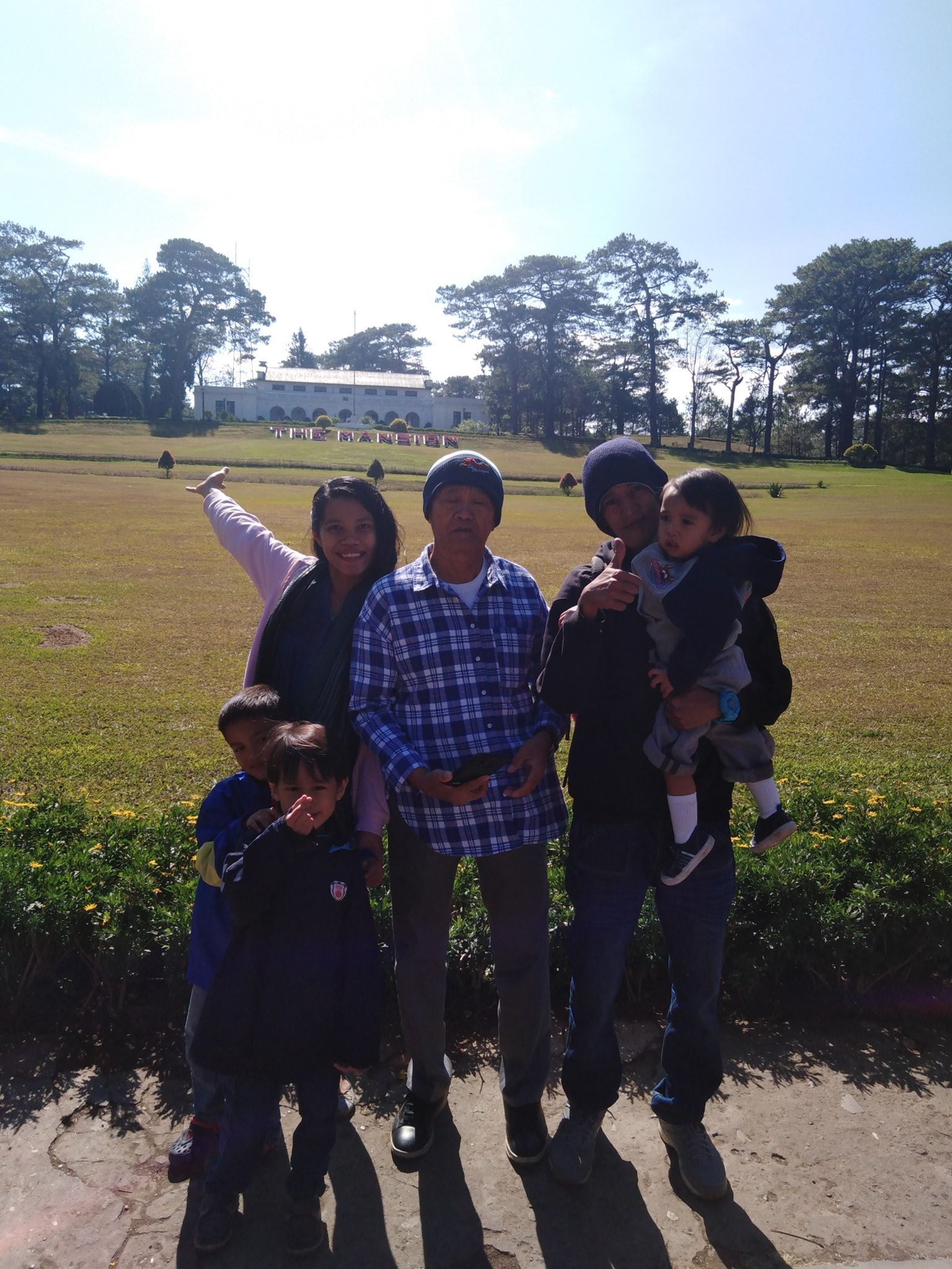 Family posing outdoors with a building in the background. Sunny day, green grass, and trees.