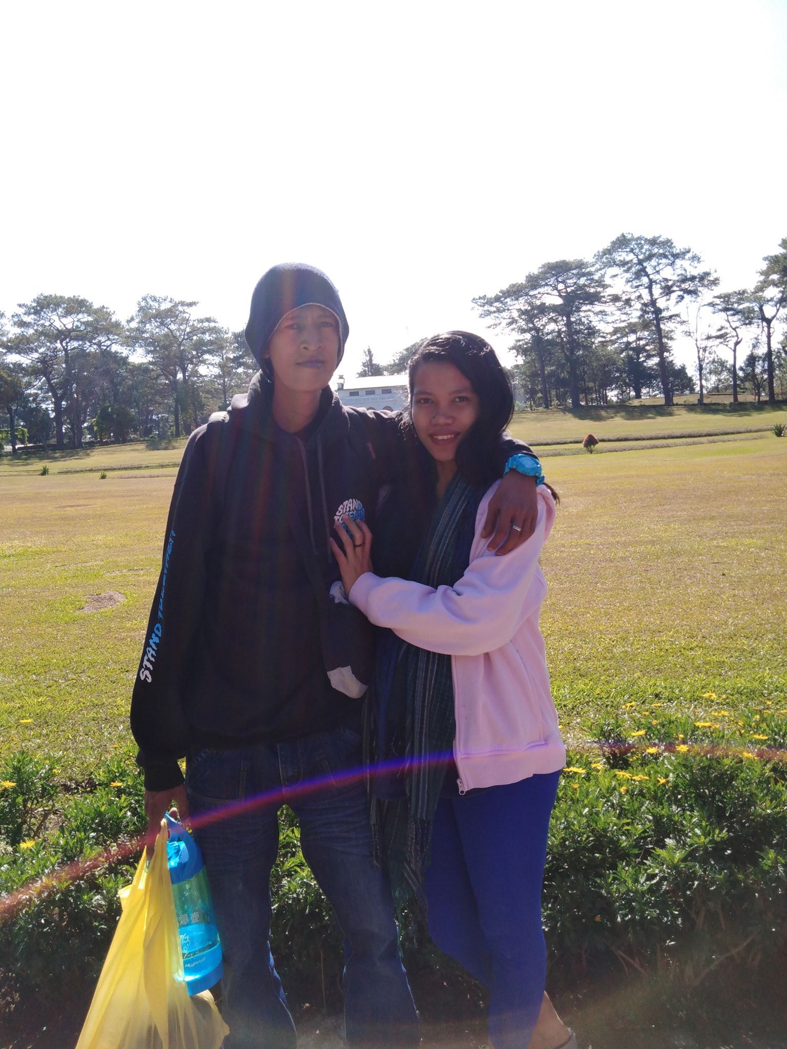 Two people outdoors; one with arm around the other. Both smiling; field and trees in the background. Sunny.