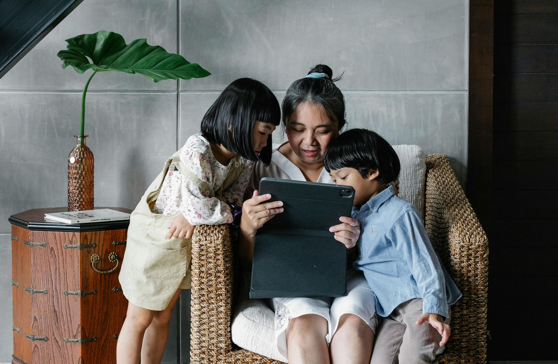 Woman and two children look at a tablet together while sitting in a wicker chair indoors.