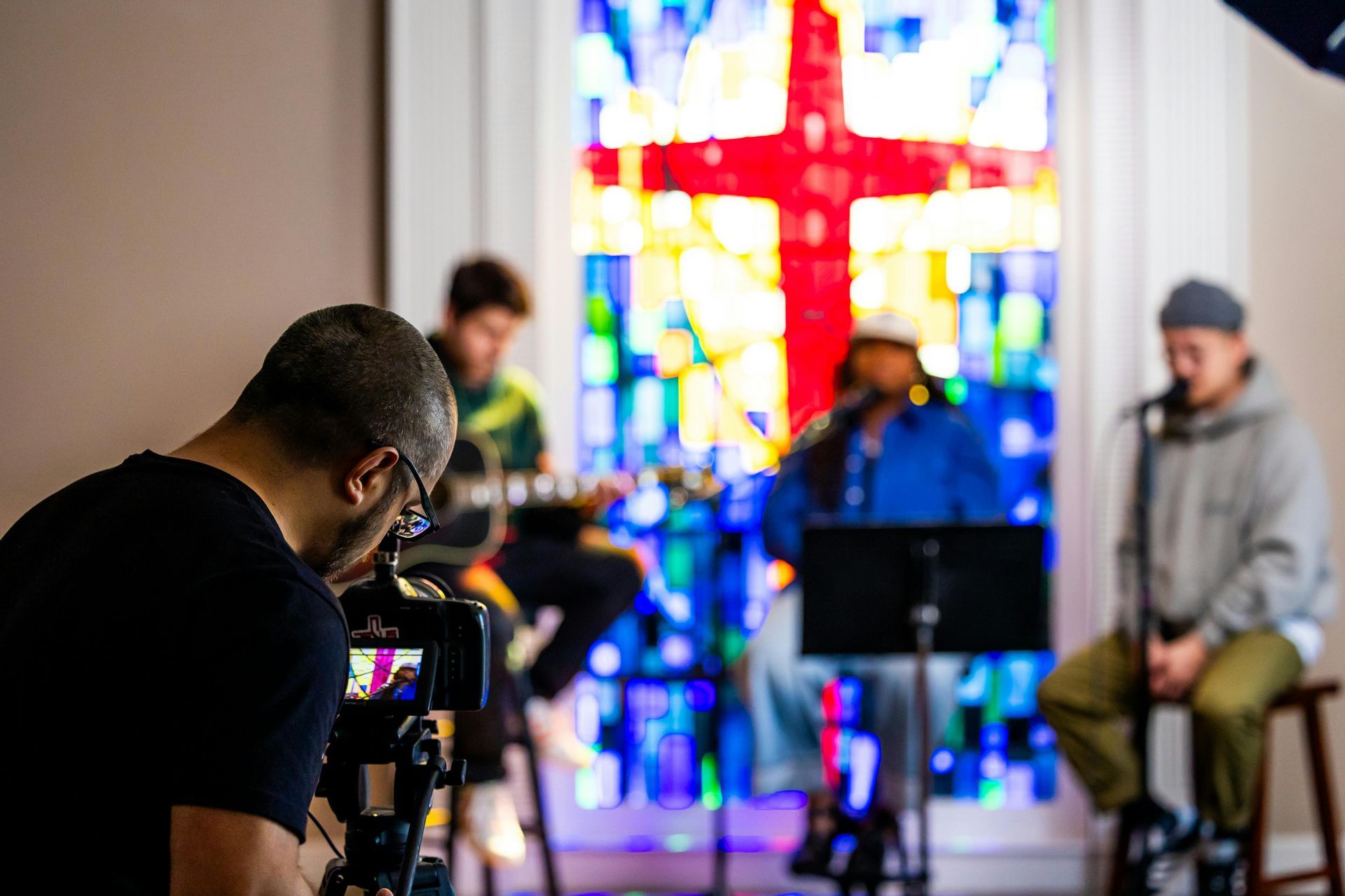 A person films a band performing in front of a stained glass window with a red cross.
