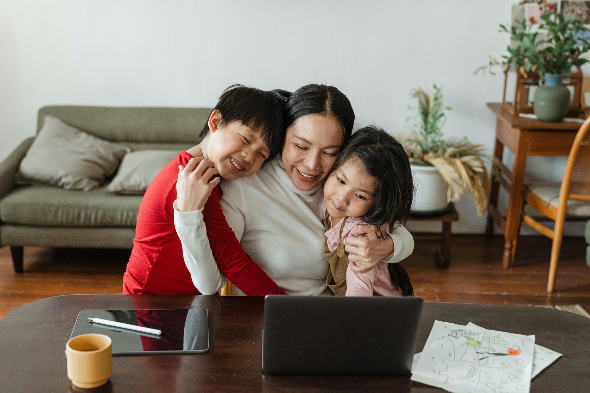 Woman embracing two children in front of a laptop at a table in a living room.