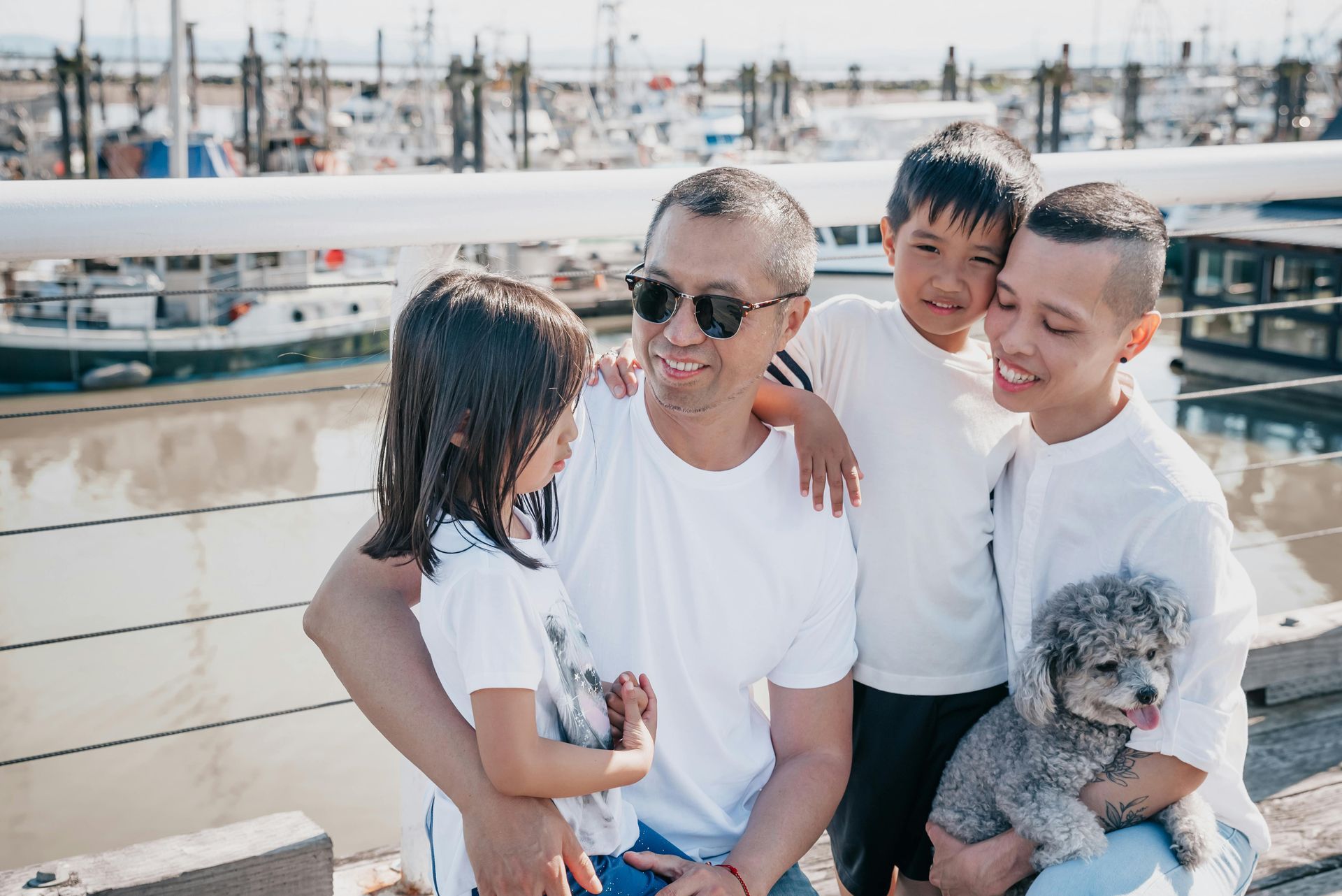 Family on dock smiling, two children and small dog. Harbor and boats in background.