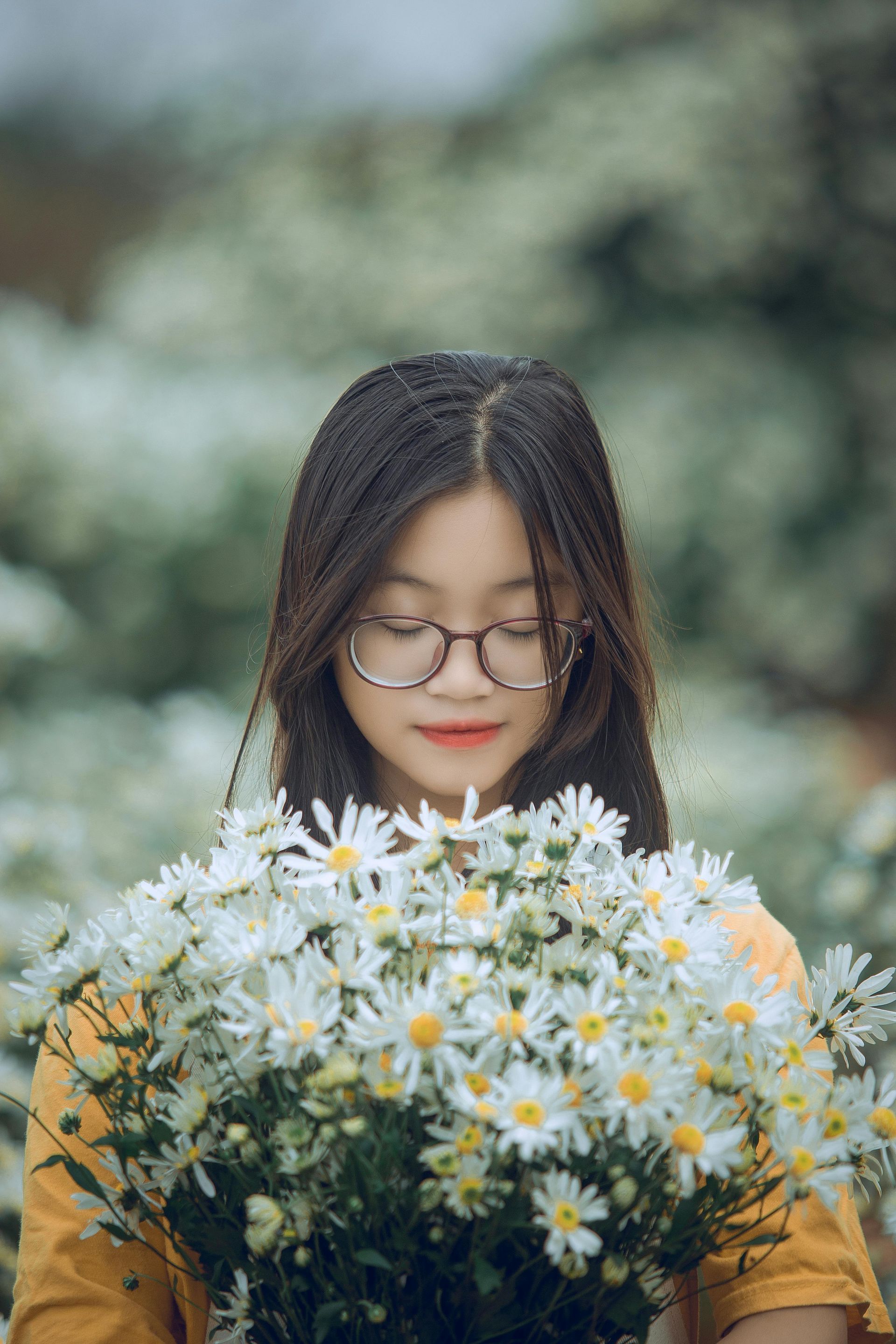 Woman holding a bouquet of white flowers, wearing glasses and a yellow shirt, surrounded by flowers.