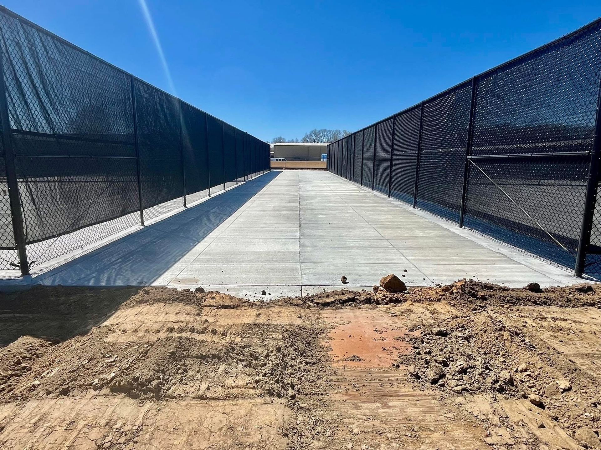 A concrete pathway stretches between two black chain-link fences under a clear blue sky, viewed from a dirt-covered foreground.