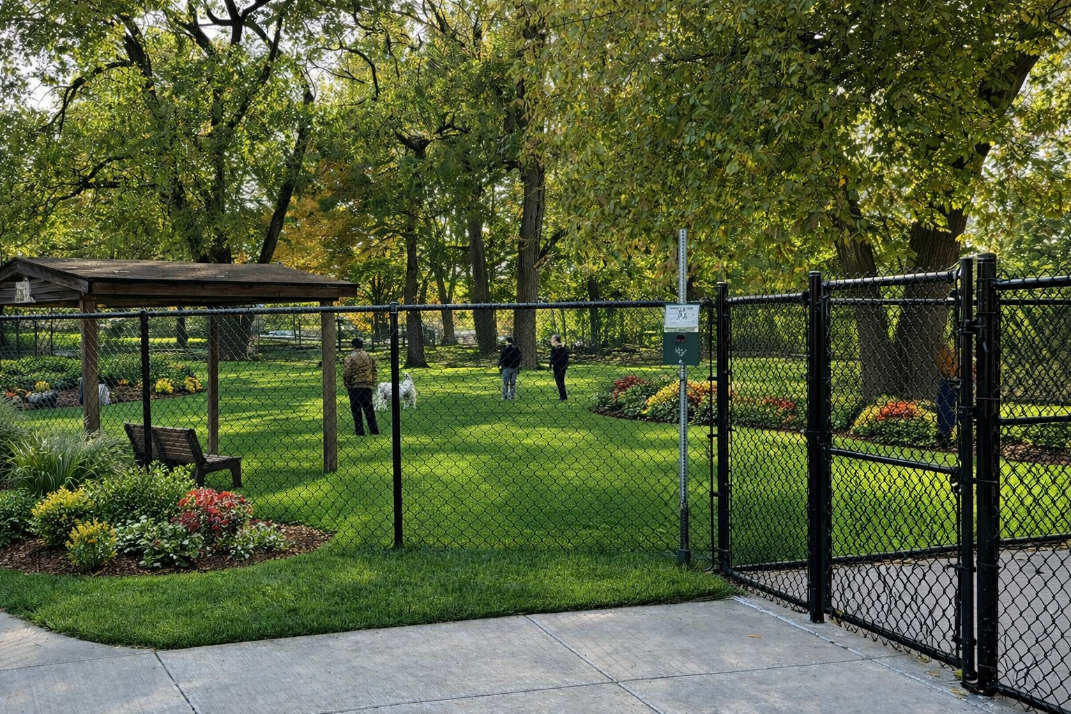 A fenced dog park with a wooden shelter, benches, green lawn, and people walking in a wooded area.