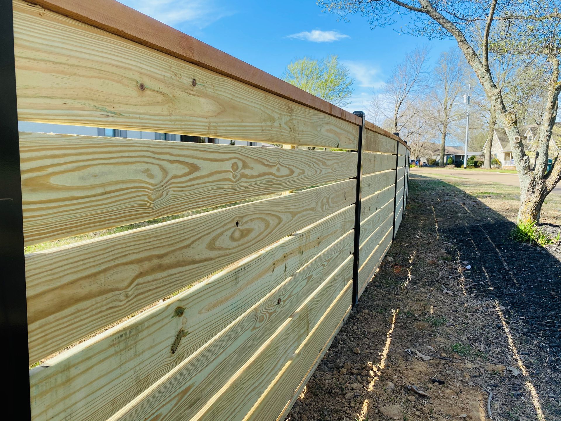 Wooden fence with horizontal slats, brown cap, and black posts along a dirt path, sunny day.