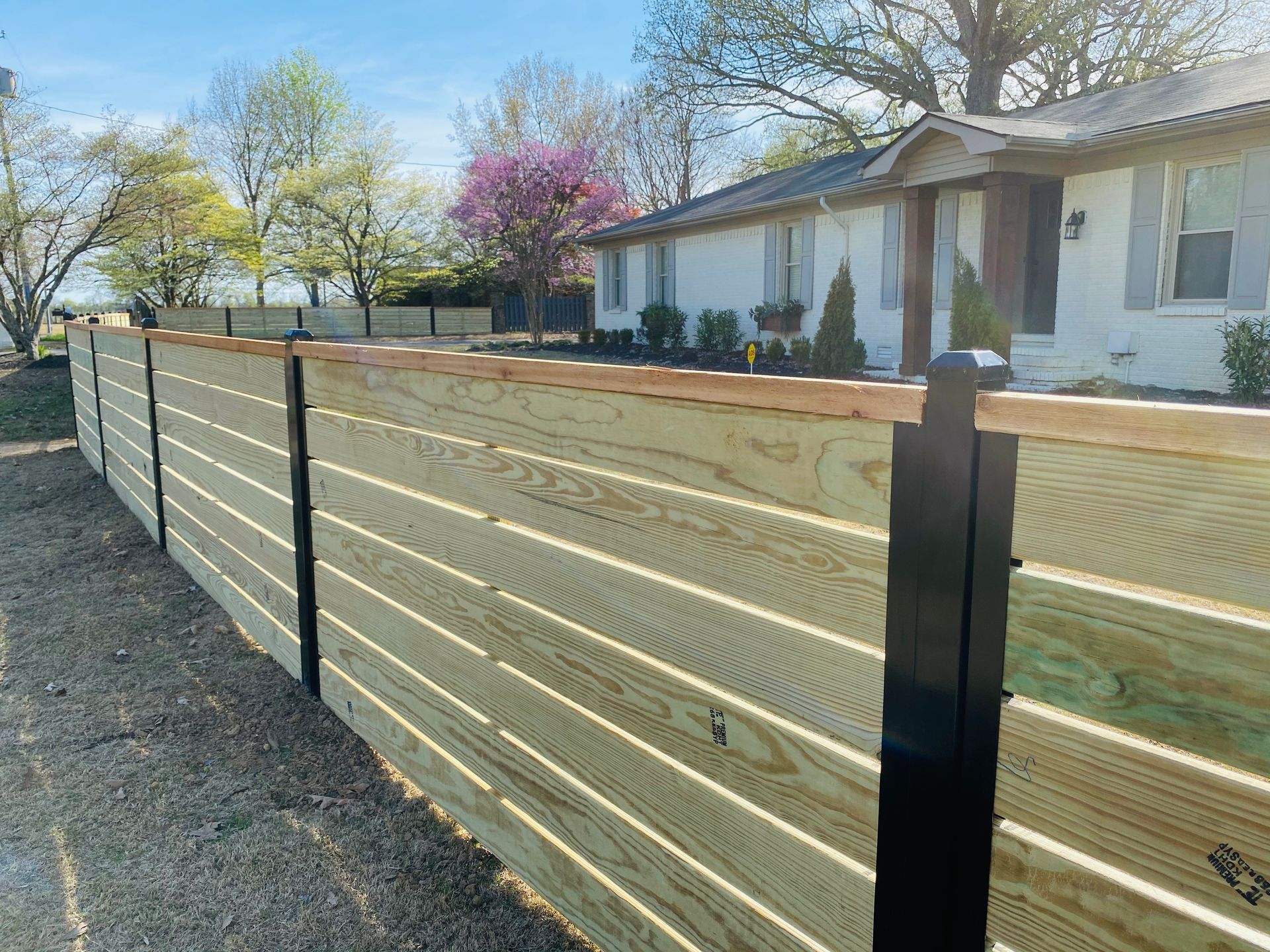 Wooden horizontal slat fence with black posts in front of a light-colored house.