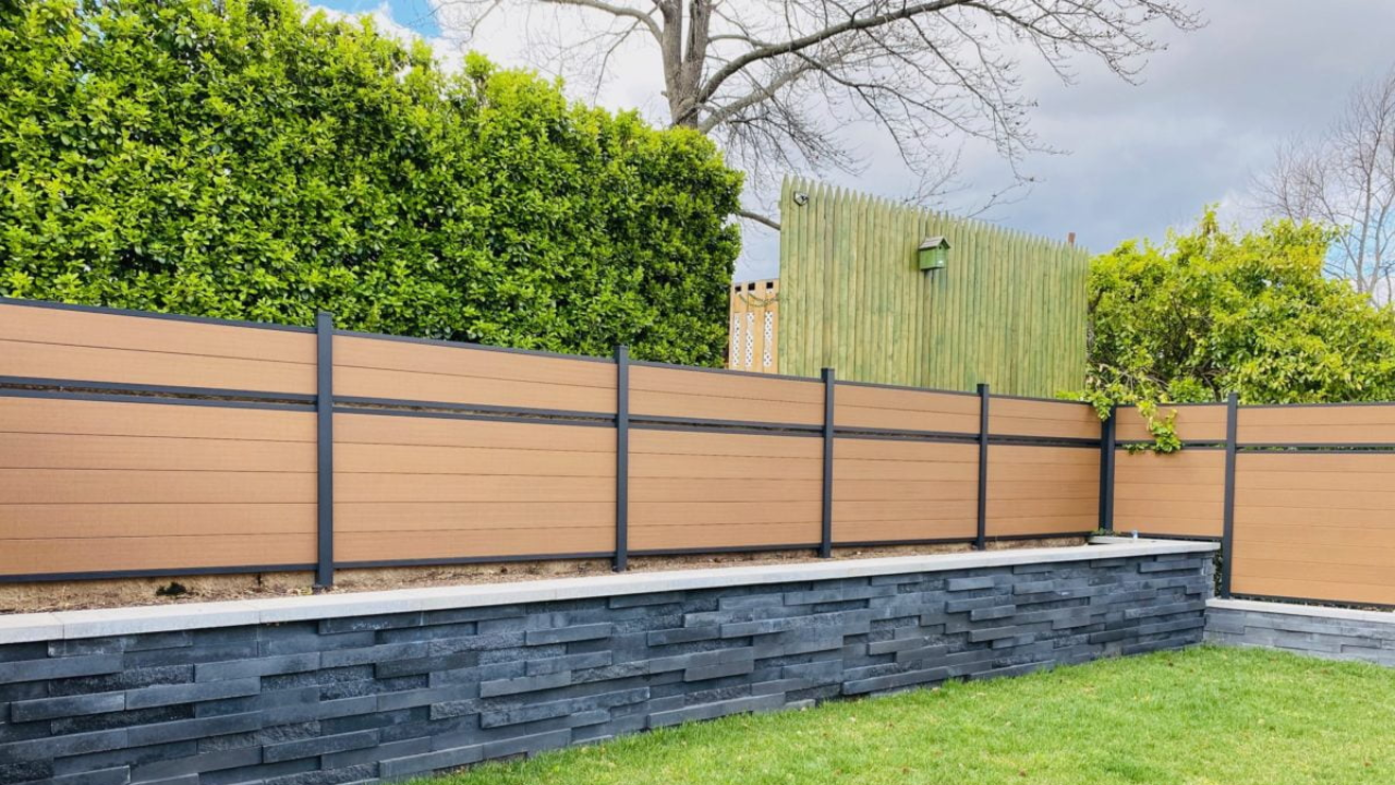 A brown and black composite fence atop a dark stone wall, with green foliage in background.