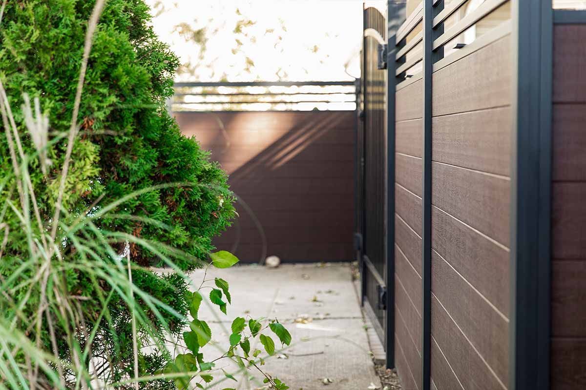 Brown wooden fence next to a green bush, with a walkway.