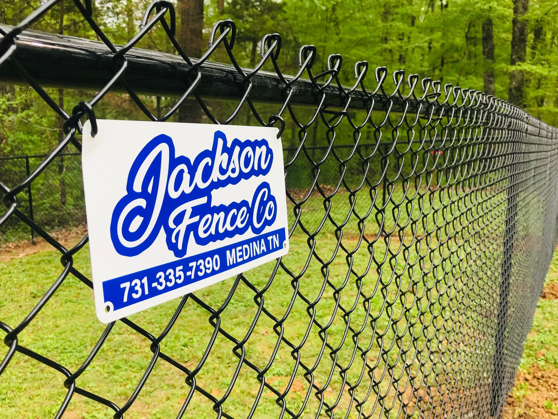 Black chain-link fence with a white sign for Jackson Fence Co. in Medina, TN; phone number visible.