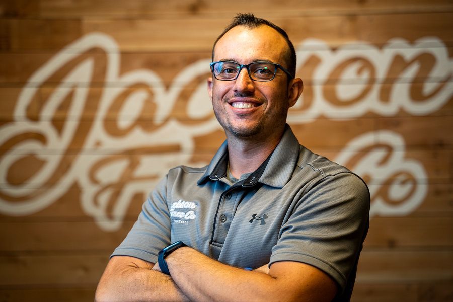 A man wearing glasses is standing with his arms crossed in front of a wooden wall.