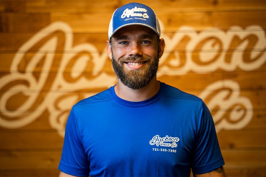 A man with a beard wearing a blue shirt and hat is smiling in front of a wooden wall.