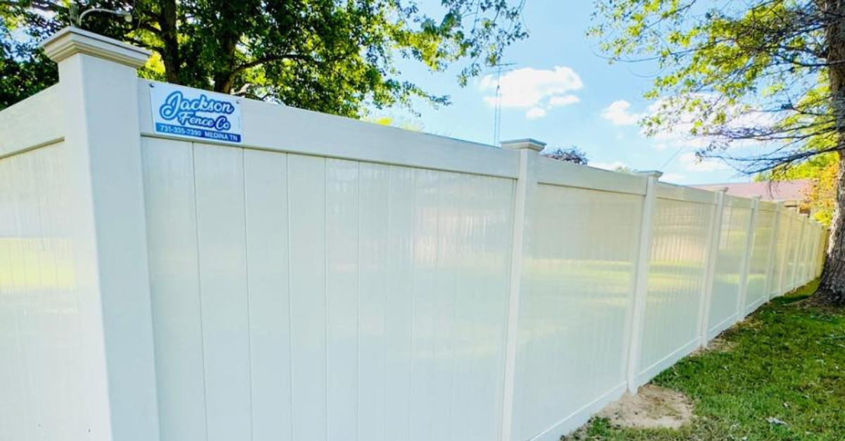 White vinyl fence along green grass, under blue sky with trees.