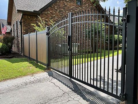 A black metal gate is open to a driveway in front of a brick house.