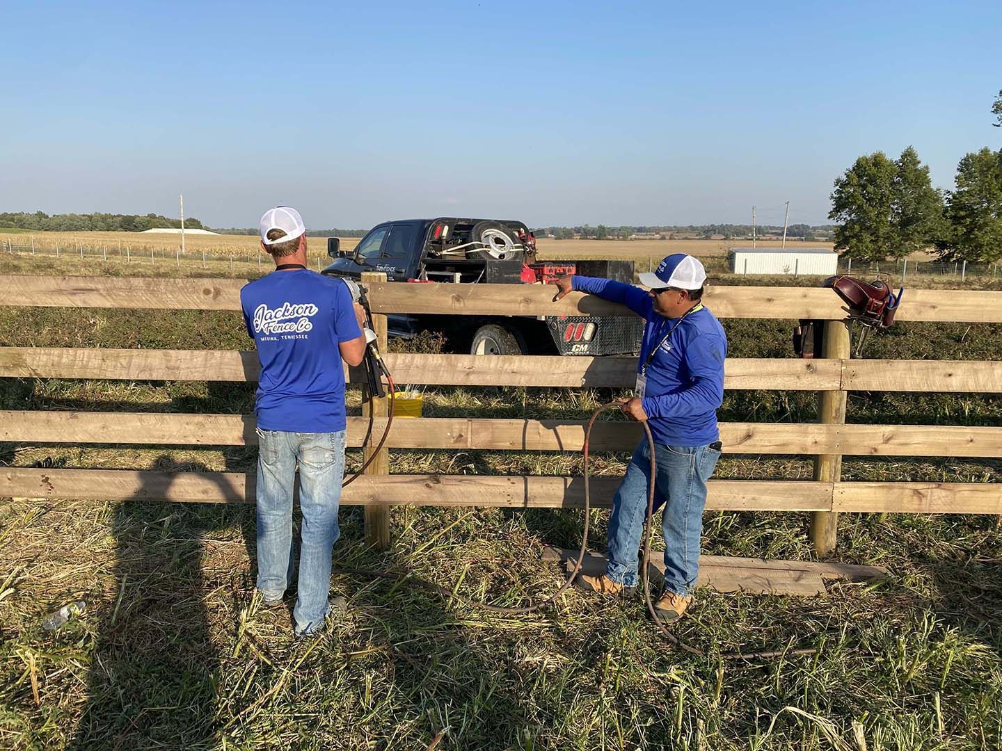 Two men are standing next to a wooden fence in a field.