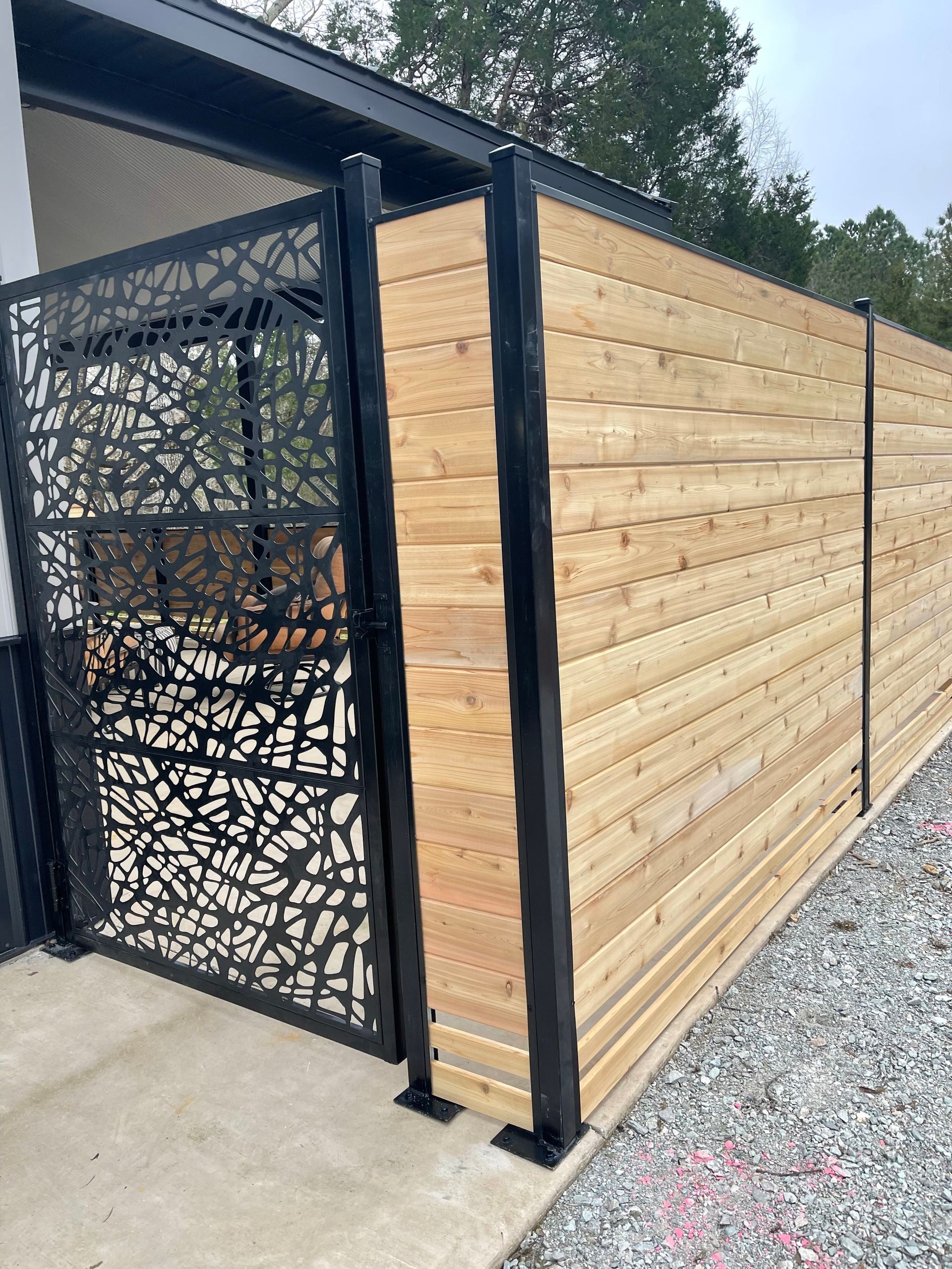 Black metal gate with a laser-cut design and a cedar plank fence, beside a building.