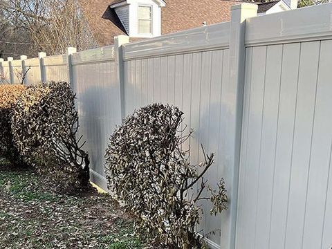 A white vinyl fence is surrounded by bushes and a house.