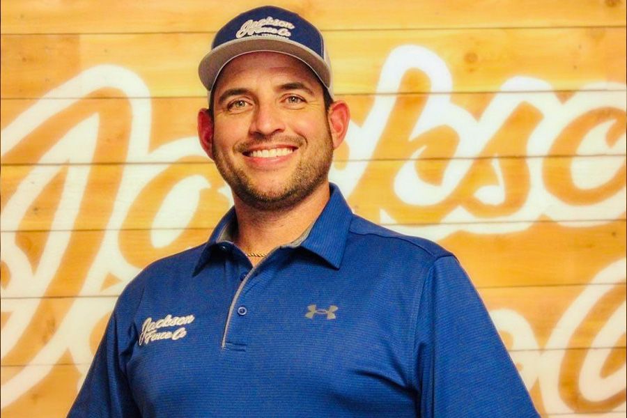 A man wearing a blue shirt and a hat is smiling in front of a sign that says welcome