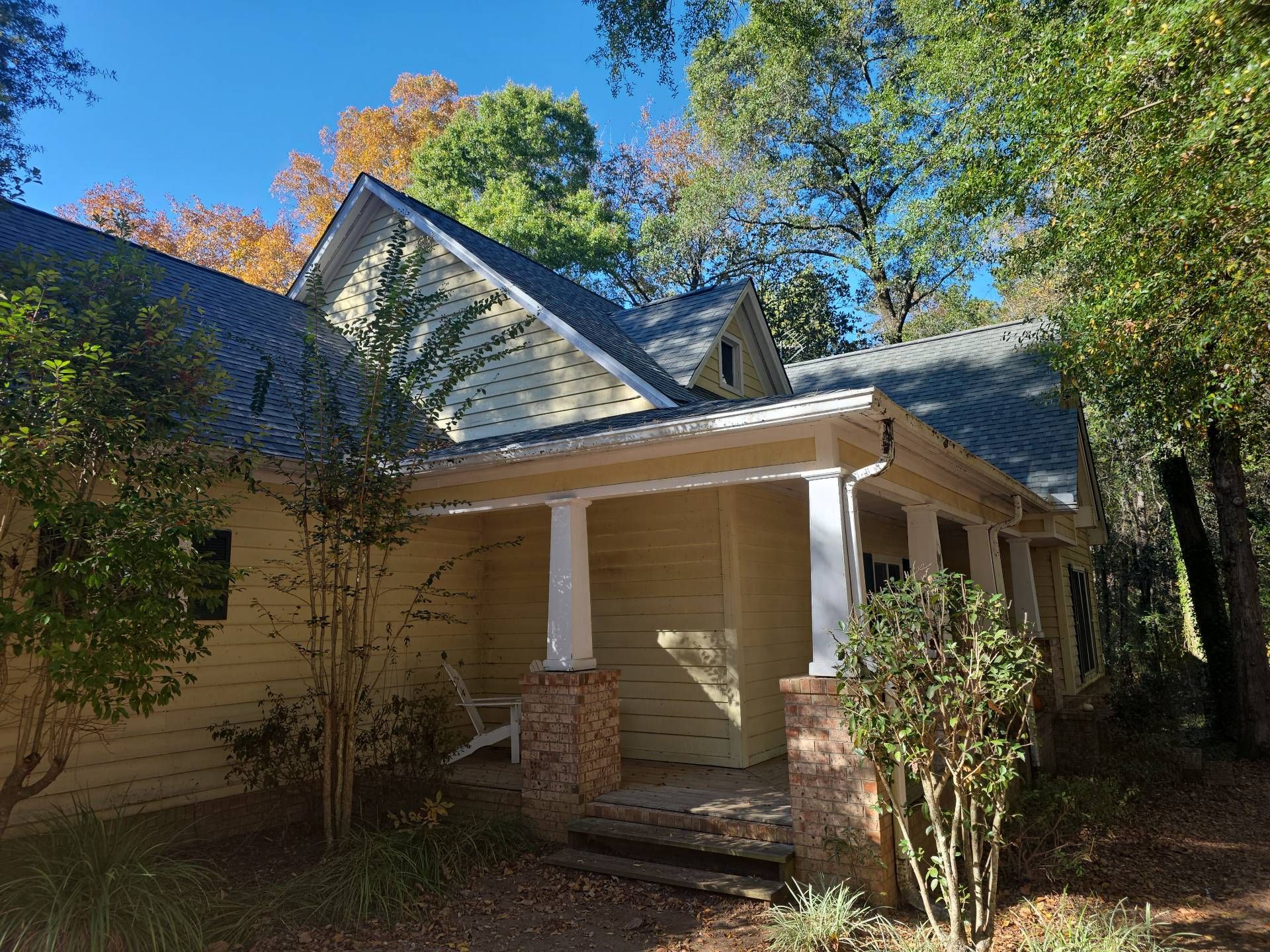 Yellow house with porch, brick columns, and a dark roof; fall foliage in the background.