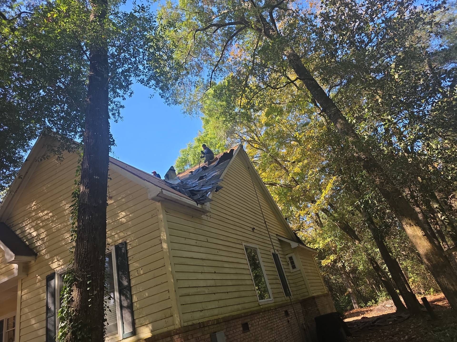 Damaged house roof against a backdrop of trees and blue sky.