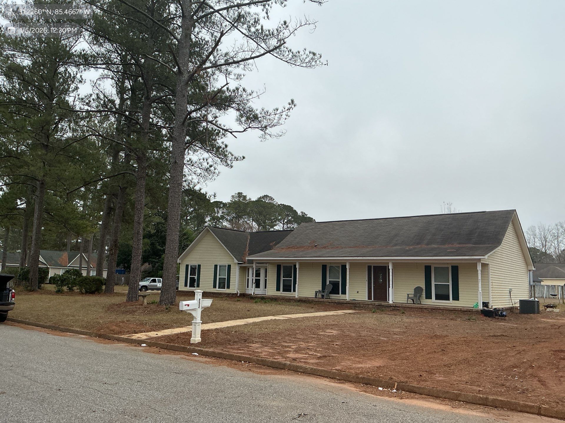 Beige ranch-style house with green shutters, brown roof, and mailbox on a cloudy day.