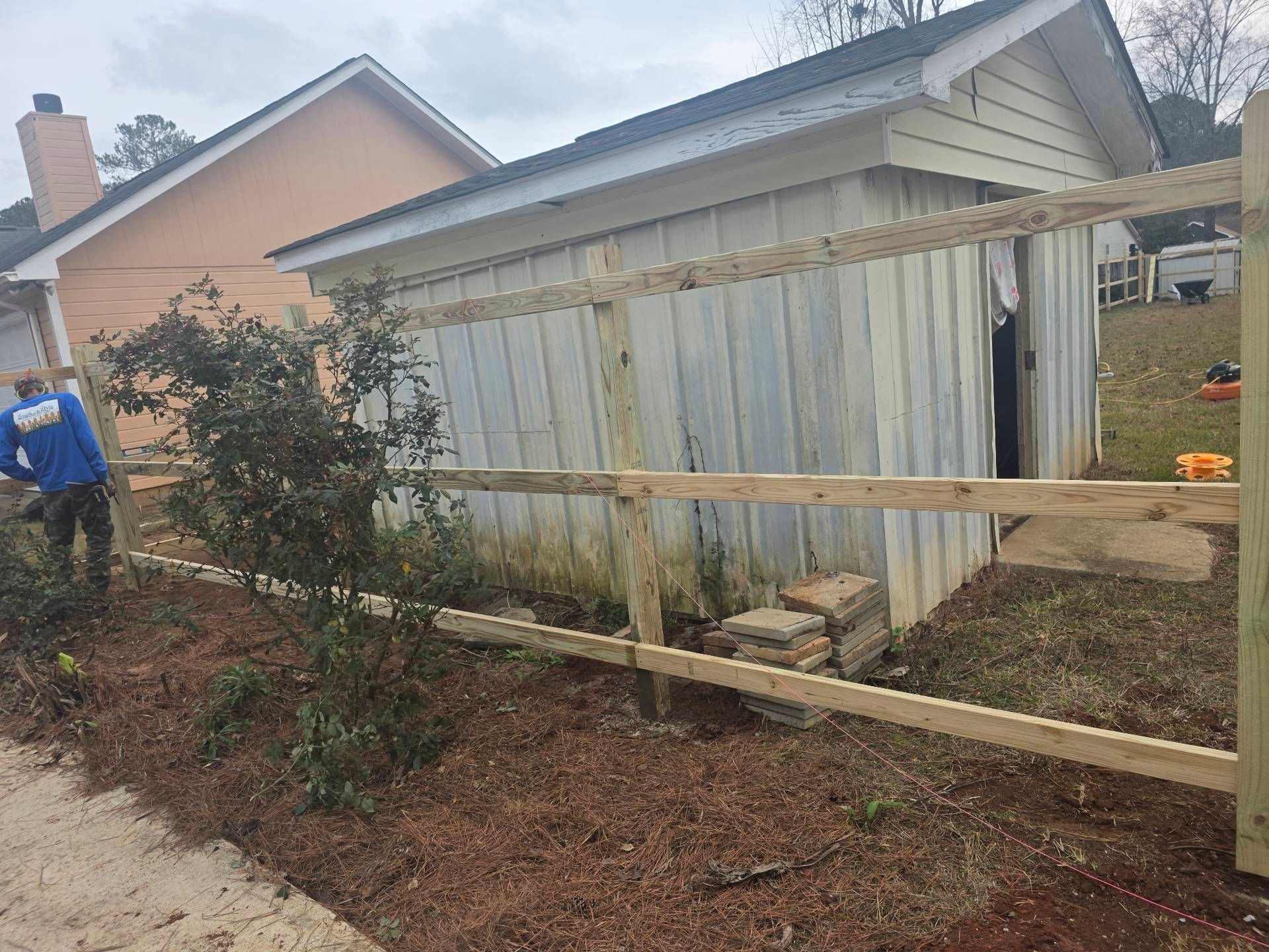 A man builds a wooden fence next to a metal shed and a house with a peach exterior.